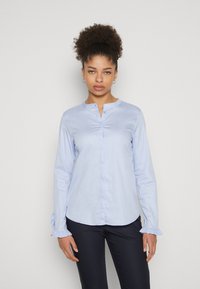 Camicia a maniche lunghe di colore blu chiaro con collo alto e chiusura a bottoni. Presenta polsini arricciati e un tessuto liscio e testurizzato.