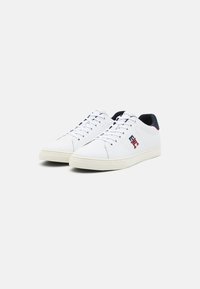Zapatillas de cuero blancas con un diseño de caña baja, que presentan un acento en azul marino y rojo en el talón y una suela de goma texturizada.