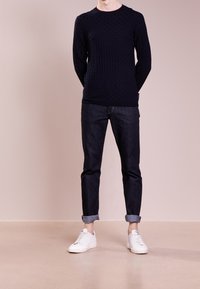 Homme portant un pull en texture sombre, un jean foncé retroussé et des baskets blanches, se tenant devant un fond rose pâle.