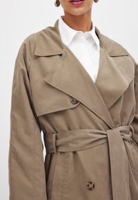Bežový trenčkot s širokým golierom, dvojradové gombíkové zapínanie a látkový opasok. Hladká textúra s voľným strihom, s bočnými vreckami.