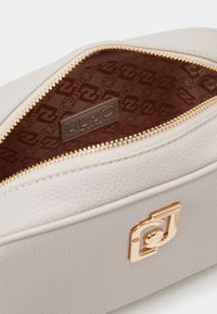 Světle šedá kožen�á taška s texturovaným povrchem, zlatým zipem a podšívkou s monogramem; logo značky viditelné uvnitř.