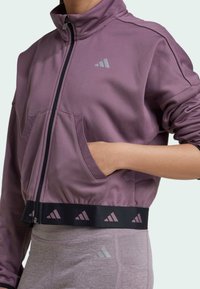 Chaqueta morada con cierre y cuello alto, y bolsillos frontales. Presenta una banda negra con acentos de logo en la cintura. Textura de tela suave.