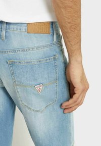Jeans in denim azzurro chiaro presentano un'etichetta triangolare del logo Guess sulla tasca posteriore, cuciture visibili e una texture liscia.