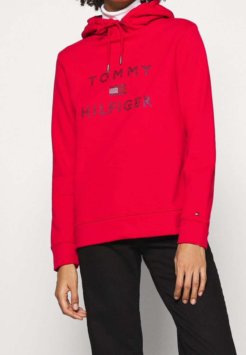 Felpa rossa con cappuccio che presenta il testo "TOMMY HILFIGER" in strass. Ha un cappuccio con coulisse e polsini a coste, abbinata a pantaloni neri.