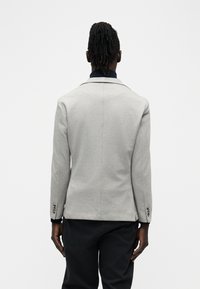 Άτομο με σκούρο δέρμα και dreadlocks, που φοράει ανοιχτό γκρι σακάκι και μαύρο παντελόνι, στεκόμενο με την πλάτη προς την κάμερα μπροστά σε λευκό φόντο.