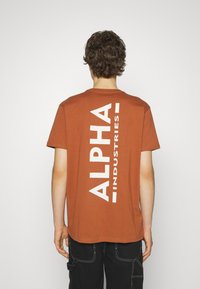 Brun bomulls t-skjorte med korte ermer, med stor hvit tekst som leser "ALPHA INDUSTRIES" vertikalt på baksiden.