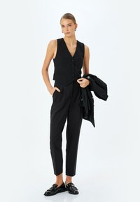 Zwarte op maat gemaakte gilet met knopen, gecombineerd met hoge zwarte pantalons. Accessoires zijn een zwart jasje en glanzende zwarte instapschoenen.