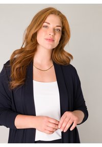 Jonge vrouw met lang rood haar draagt een zwarte cardigan, witte top en gouden ketting, staand tegen een effen lichte achtergrond.