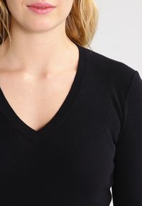 Femme à la peau claire et aux cheveux blonds portant un pull noir en maille côtelée avec un décolleté en V, montrée des épaules aux lèvres sur un fond uni.