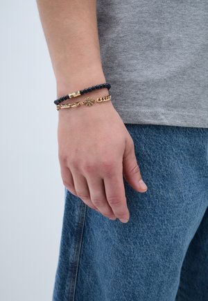 Juodų karoliukų apyrankė ir auksinė grandinėlė su gėlių akcentais, nešiojama ant rankos prieš pilką marškinėlį ir mėlynus džinsus.