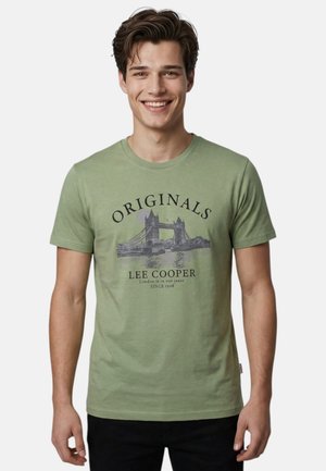 Jeune homme souriant portant un T-shirt vert clair avec un graphisme du Tower Bridge et le texte "ORIGINALS LEE COOPER London est dans nos jeans DEPUIS 1908".