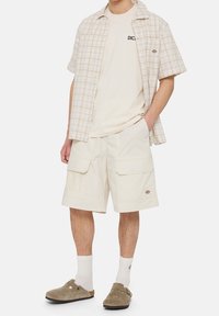 Mies, jolla on yllään beiget cargo-shortsit, valkoiset sukat, taupe-väriset liukuvaatteet, kerma-valkoinen t-paita ja auki oleva lyhythihainen ruudullinen paita, seisoo.