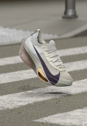Ανοιχτόχρωμο αθλητικό παπούτσι Nike με σκουρόχρωμο σήμα που αιωρείται πάνω από διάβαση πεζών σε πλακόστρωτο δρόμο.