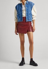 Gilet in denim di colore blu, camicia beige, gonna mini in pelle rossa e scarpe nere. Caratteristiche includono un design corto e bottoni visibili.