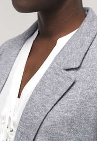 Grå blazer med texturerad tyg, med en notched krage och en ren design. Bärs över en vit snörningstopp.