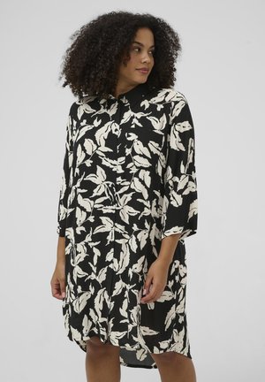 Kvinde med krøllet hår iført en sort skjortekjole med hvide bladmønstre, stående og kiggende mod sin højre side foran en ensfarvet baggrund.