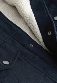 Navy wool jacket featuring a soft cream shearling lining, metal buttons, and patch pockets. Textured exterior with a structured design.