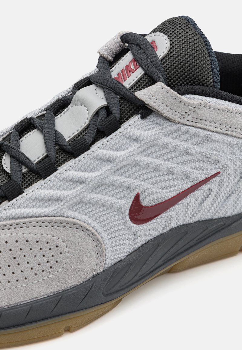 Nike-Sneaker aus grauem Stoff mit strukturierten Wildlederakzenten. Merkmale sind der burgunderfarbene Swoosh, die graue Mesh-Zunge und schwarze Schnürsenkel. Gummisohle.