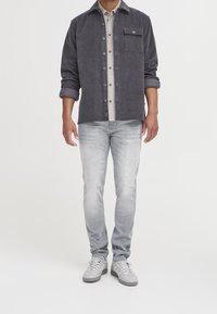 Chemise en velours côtelé gris avec une fermeture à boutons, dotée d'une poche poitrine, portée sur une chemise à carreaux claire ; associée à un jean en denim clair et à des baskets grises.