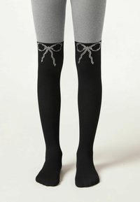Sorte strømpebukser med en satinlignende finish, prydet med et dekorativt buebånddesign i rhinsten ved knæet, parret med grå leggings ovenfor.