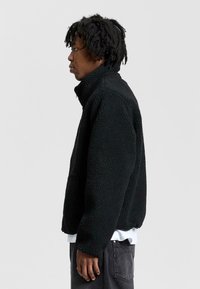 Musta fleece-takki korkealla kauluksella, teksturoidulla pinnalla ja rento istuvuus. Sisältää etutaskun ja on kerroksittain valkoisen paidan päällä.