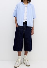 Camicia a maniche corte a righe blu chiaro, canotta bianca, pantaloni corti a gamba larga in denim scuro, sneakers bianche con dettagli grigi e calze a lunghezza media.