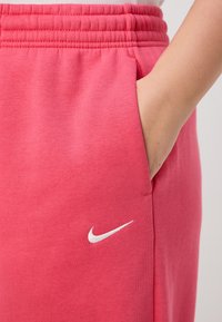 Rožinės medvilnės kelnes su elastinga juosta ir šonine kišene, su balta Nike logotipu apatiniame kairiajame kampelyje. Lygus paviršius.