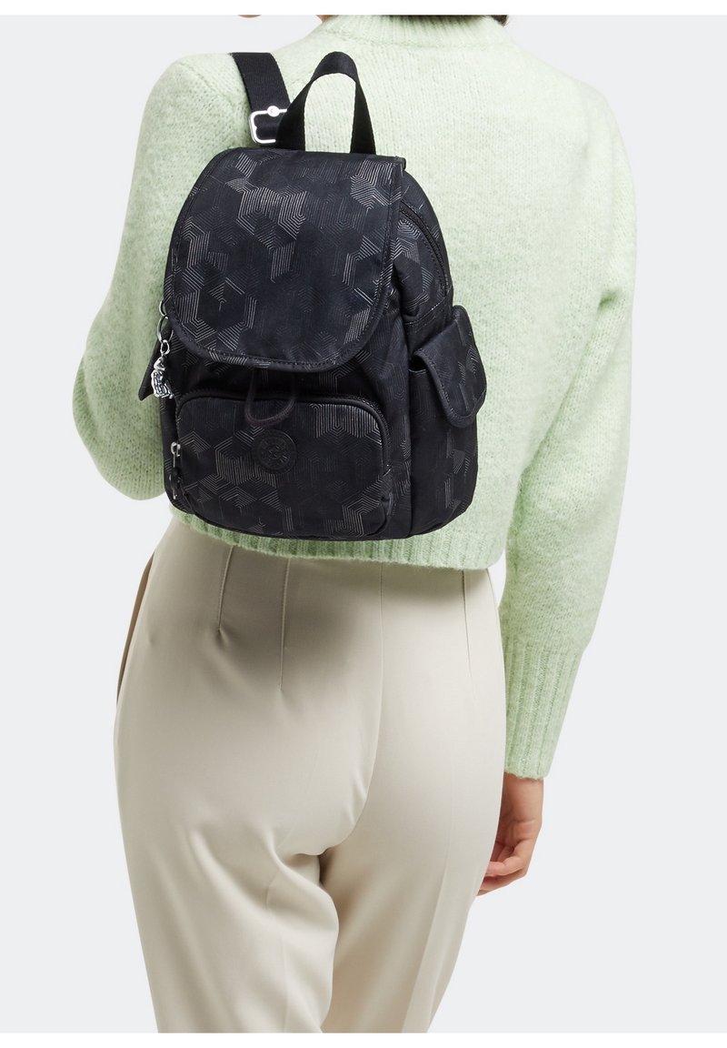 Personne portant un pull vert clair et un pantalon beige, portant un sac à dos noir à motifs avec des poches avant et latérales.