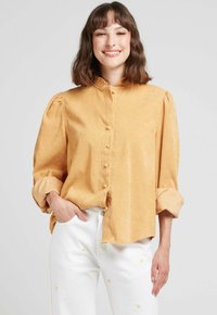 Blouse jaune moutarde à manches longues avec un col froncé et une fermeture à boutons. Texture douce ; accompagnée de pantalons blancs ornés de broderies florales.