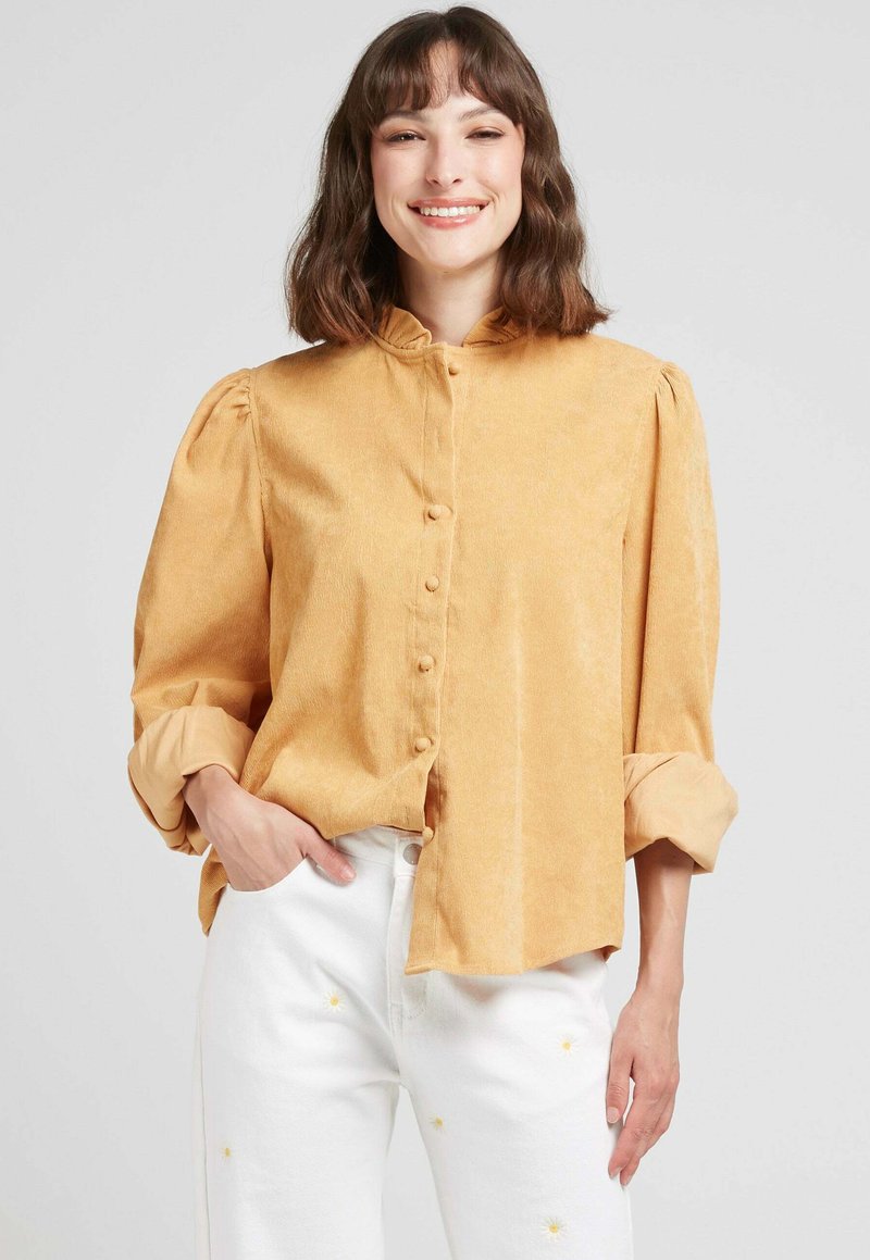Blouse jaune moutarde à manches longues avec un col froncé et une fermeture à boutons. Texture douce ; accompagnée de pantalons blancs ornés de broderies florales.