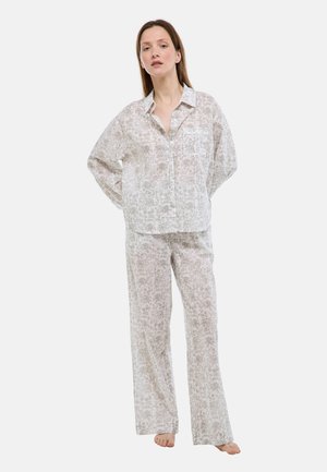 Femme debout, pieds nus, portant un ensemble de pyjama ample à manches longues de couleur claire avec un motif floral, composé d'une chemise boutonnée et d'un pantalon à jambes larges.