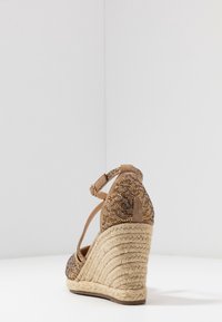Sandalia de cuña con tacón de yute trenzado. La parte superior está hecha de ante marrón y presenta un diseño texturizado con cuentas. Correa ajustable en el tobillo para el cierre.
