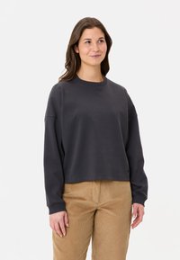 Felpa nera corta con collo tondo ampio e spalle cadenti, realizzata in tessuto liscio. Indossata con pantaloni in corduroy beige.