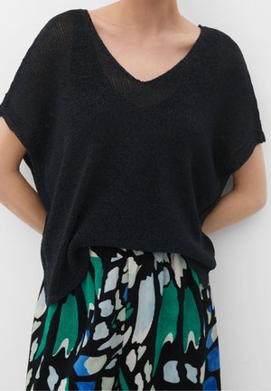 Personne portant un haut en maille noire ample à col en V, associé à une jupe plissée avec des motifs abstraits audacieux en vert, bleu, noir et blanc.