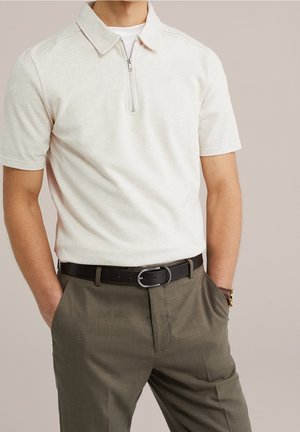 Homme portant un polo zippé beige clair à manches courtes, ceinture marron et pantalon vert olive, les mains dans les poches sur un fond neutre.