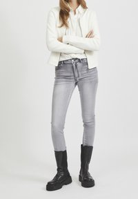 Vit vadderad bomberjacka över en ljus skjorta, i kombination med grå tajta jeans och knähöga svarta stövlar. Släta texturer, elegant design.