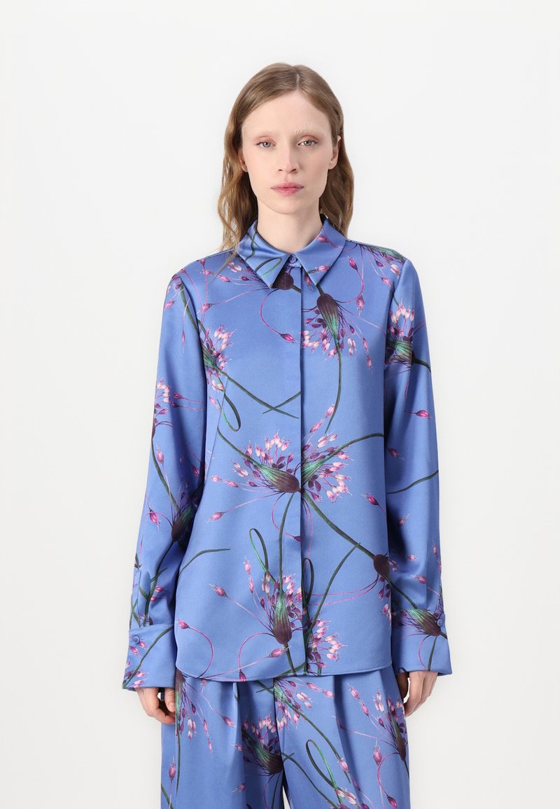 Γαλάζιο πουκάμισο με μακριά μανίκια, με εμπριμέ λουλουδιά σε μωβ και πράσινο. Διαθέτει αιχμηρό γιακά και κλείσιμο με κουμπιά στο μπροστινό μέρος.