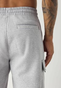 Gros plan d'une personne portant un pantalon de survêtement gris clair avec une ceinture élastique, une poche arrière et une poche cargo latérale, montrant un bras tatoué.