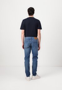 Hombre de pie de espaldas, vistiendo una camiseta negra, jeans azules y zapatillas blancas, frente a un fondo blanco liso.