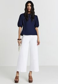 Top blu navy a maniche corte con vita arricciata, abbinato a pantaloni bianchi a gamba larga. La modella indossa scarpe con plateau marroni e tiene una borsa all'uncinetto.