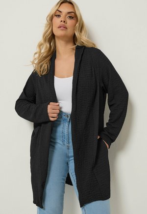 Lyshåret kvinde iført en textureret sort åben cardigan over en hvid tanktop og lyseblå jeans, stående mod en ensfarvet baggrund.