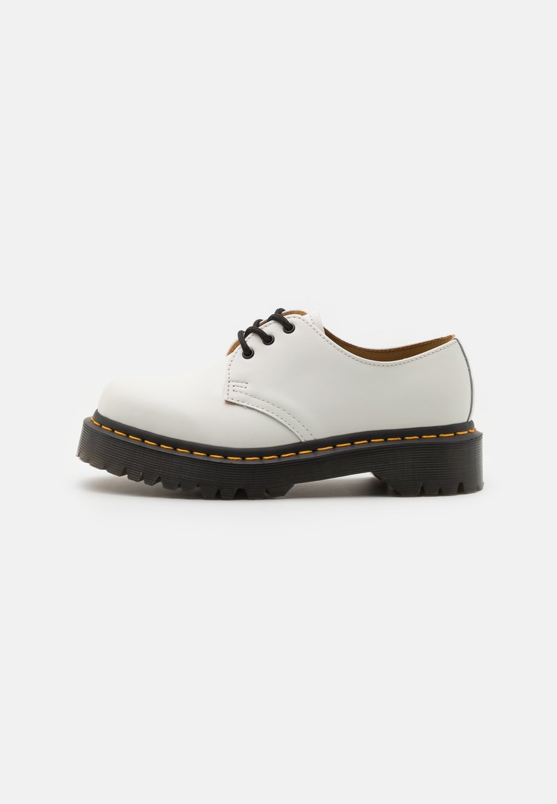 Chaussure basse en cuir blanc avec semelle épaisse noire, surpiqûres jaunes et lacets noirs, vue de côté sur fond blanc.