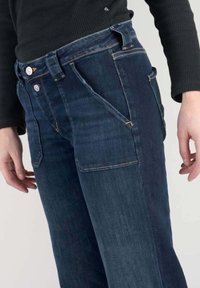 Mörkblå denim jeans med distinkta framfickor, synlig sömnad och stängning med två knappar. Tyget verkar något texturerat med en avslappnad passform.