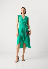 Green dress with a deep V-neck and ruffle sleeves. Features a fitted waist and asymmetric hem. Paired with a black clutch and heels.