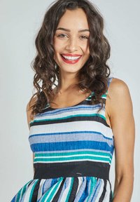 Mujer sonriente con cabello rizado oscuro, vestida con un vestido sin mangas a rayas azul, verde, negro y blanco, contra un fondo liso.