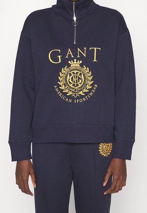 Mężczyzna w granatowej bluzie i dopasowanych spodniach z żółtym logo i emblematem GANT, bluza ma kołnierz z półzamkiem.