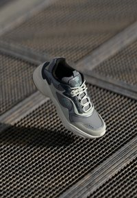 Gray sneaker with textured upper, featuring lighter gray accents, a padded collar, and reflective details. Laces are lightly crisscrossed.