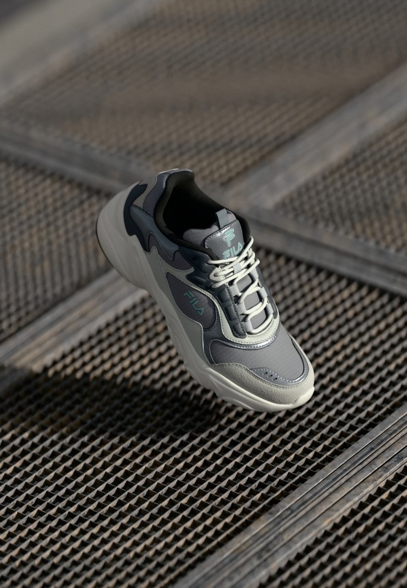 Gray sneaker with textured upper, featuring lighter gray accents, a padded collar, and reflective details. Laces are lightly crisscrossed.