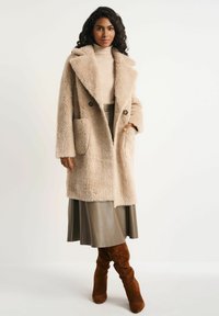 Beżowy, fakturowany płaszcz z dużym kołnierzem, na guziki, z kieszeniami. Połączony z kremowym golfem i plisowaną oliwkową spódnicą.