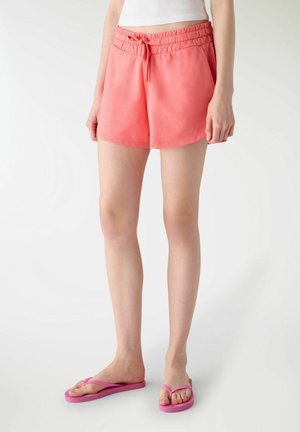 Donna che indossa pantaloncini corallo con coulisse, canotta bianca corta e infradito rosa, in piedi contro uno sfondo bianco.